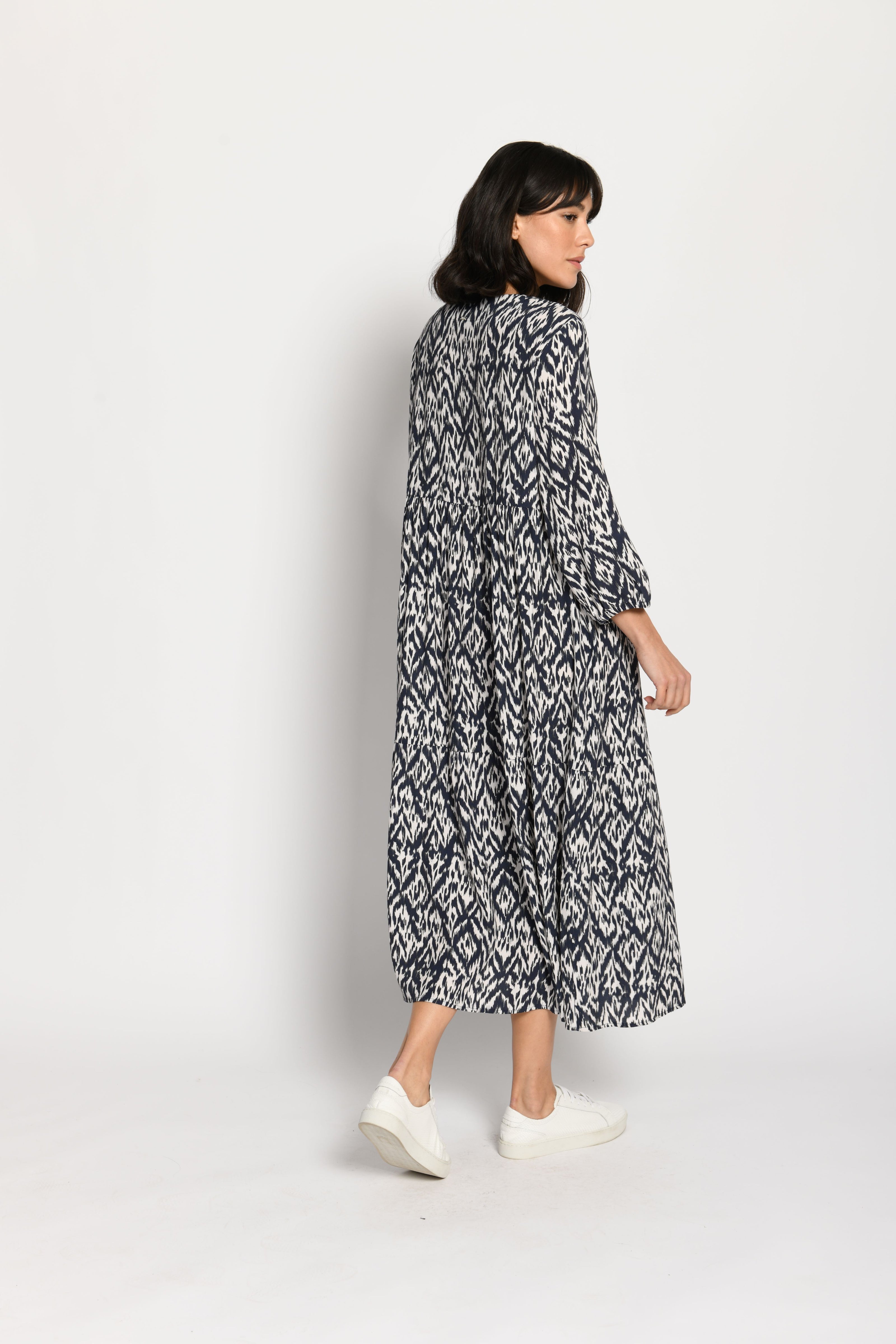 Model trägt luftiges Stufenkleid mit langen Ärmeln und tollem Print in den Farben Weiß und Dunkelblau – Rückansicht
