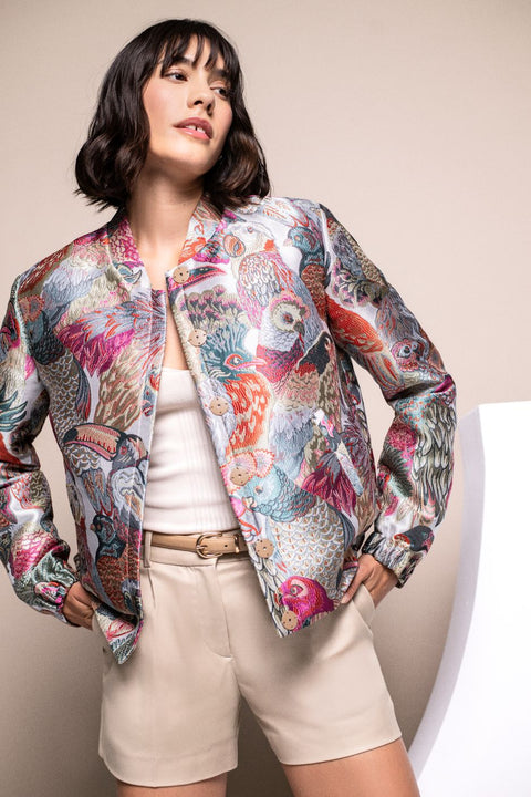 Model trägt locker geschnittenes Blouson mit buntem Vogelmuster, zartem Glanz und elastischem Ärmelbündchen.