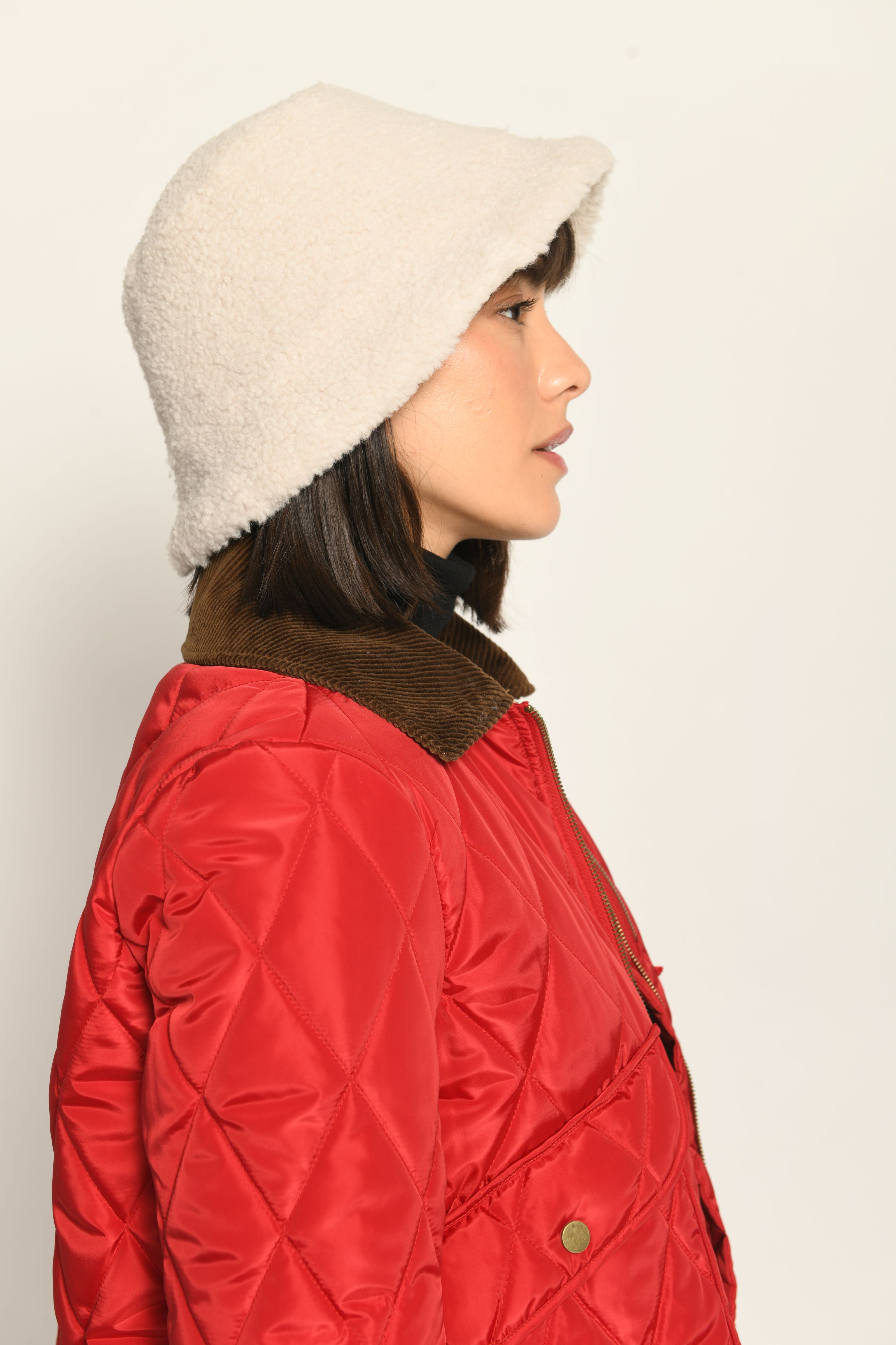 Model trägt Hut aus flauschigem cremefarbenem Teddy-Webpelz, lässig kombiniert zu einer roten Jacke – Seitenansicht