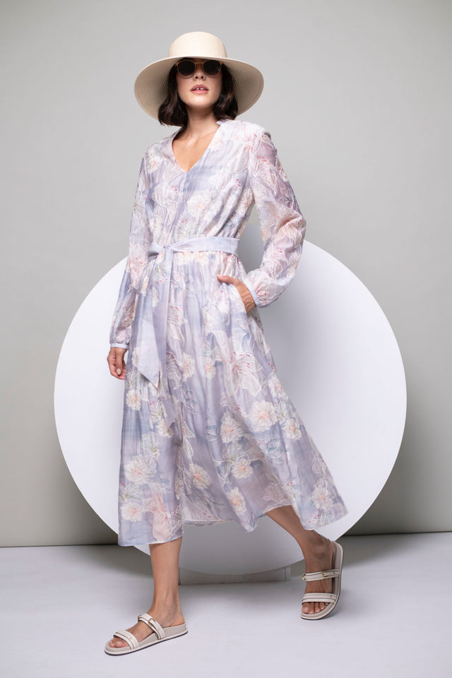 Model trägt bedrucktes, langärmeliges Kleid mit zartem Pastellprint, kombiniert mit Basthut und offenen Schuhen.