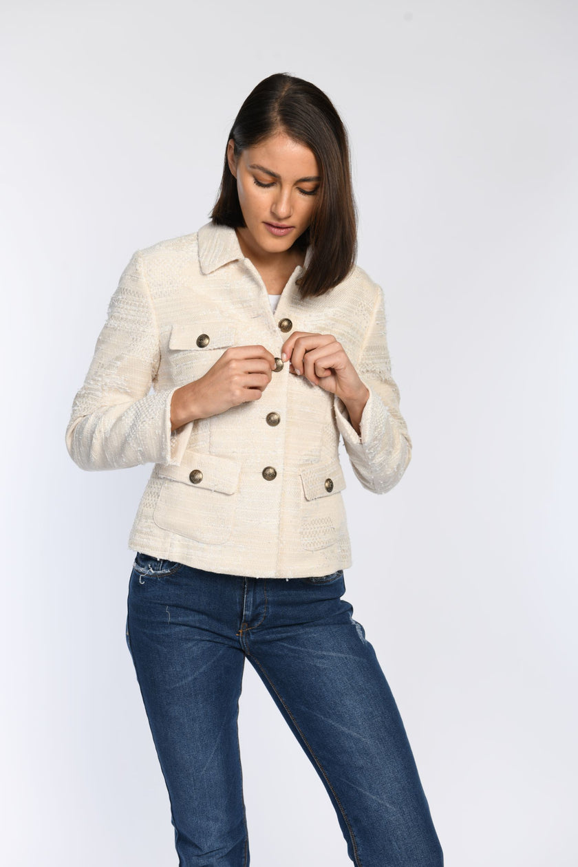 Model knöpft cremefarbene, langärmelige Jacke mit großen dekorativen Knöpfen zu, kombiniert mit blauer Jeans.