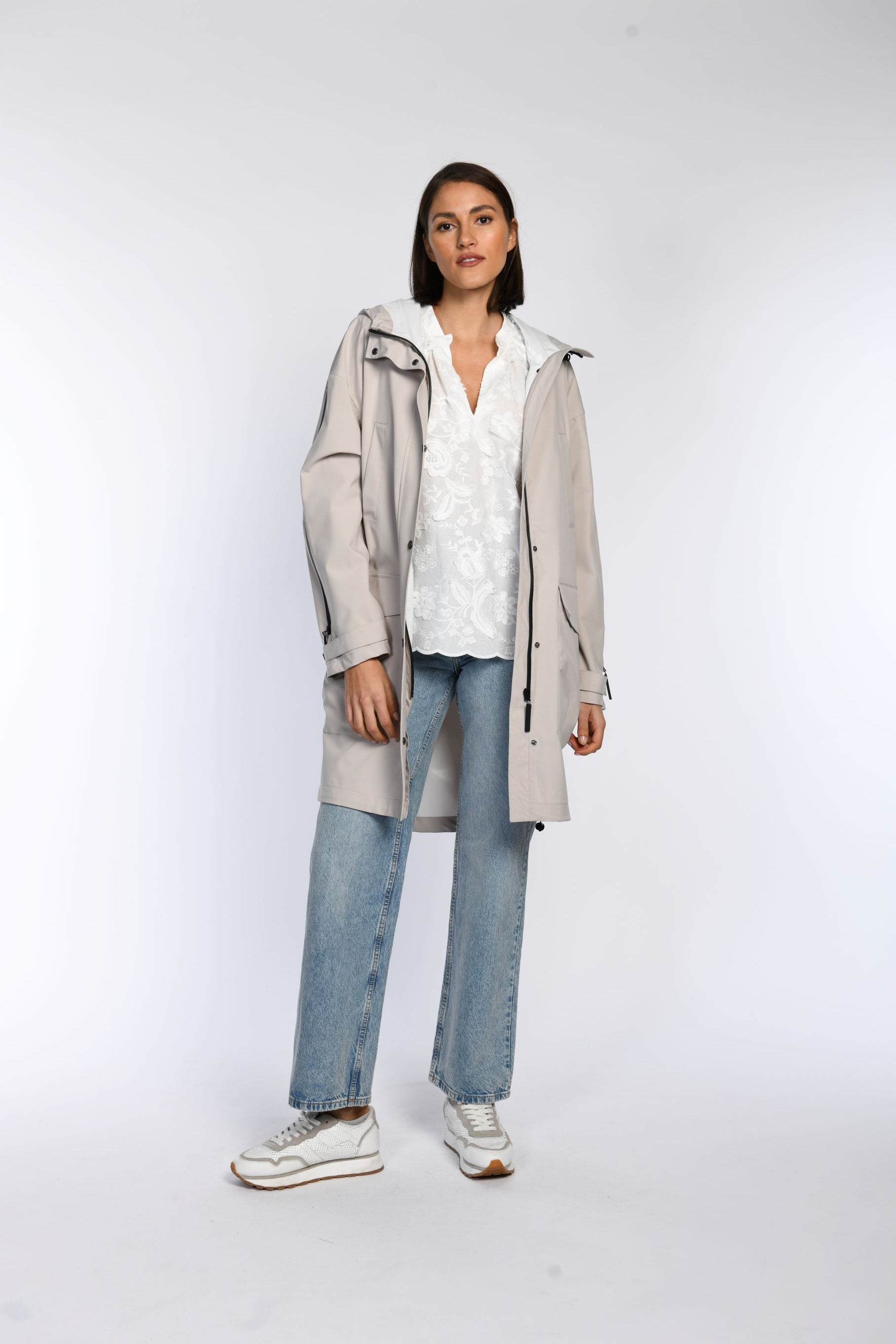 Das Model trägt einen offenen, beigen Regenparka, der mit einer weißen Bluse mit V-Ausschnitt kombiniert wird.